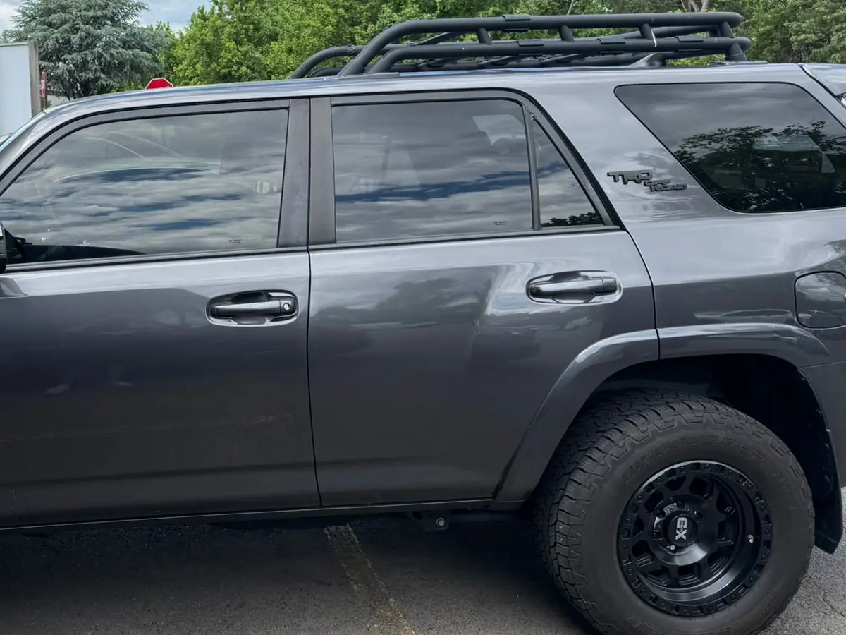 A charcoal grey Toyota 4Runner SUV with black wheels, all-terrain tires and a roof rack, parked in side profile beside a cream brick building under a bright sky.