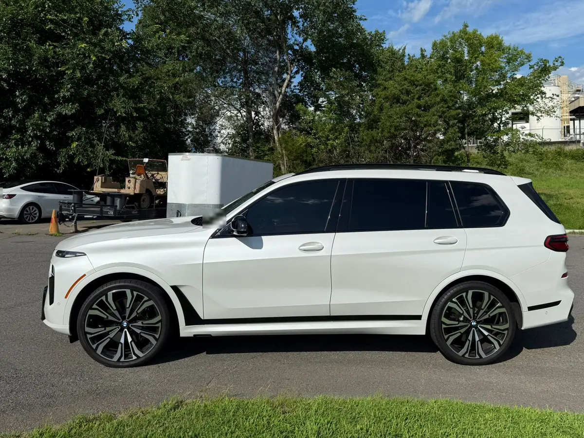 A white BMW X7 parked in side profile on an open lot, with a treeline and blue sky behind it.