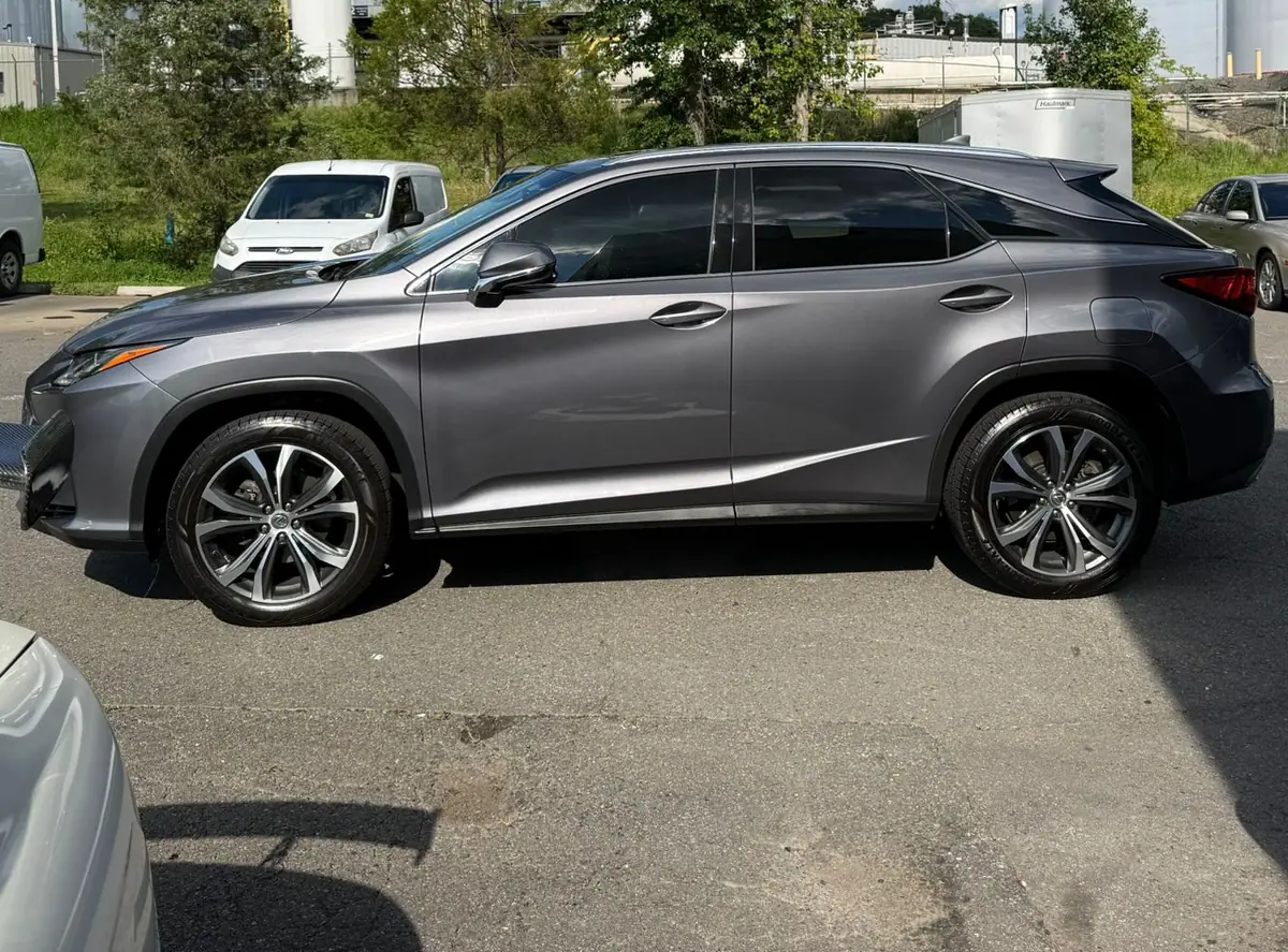 A dark grey Lexus SUV parked side-on in a sunlit lot, with vans and industrial storage tanks behind it.