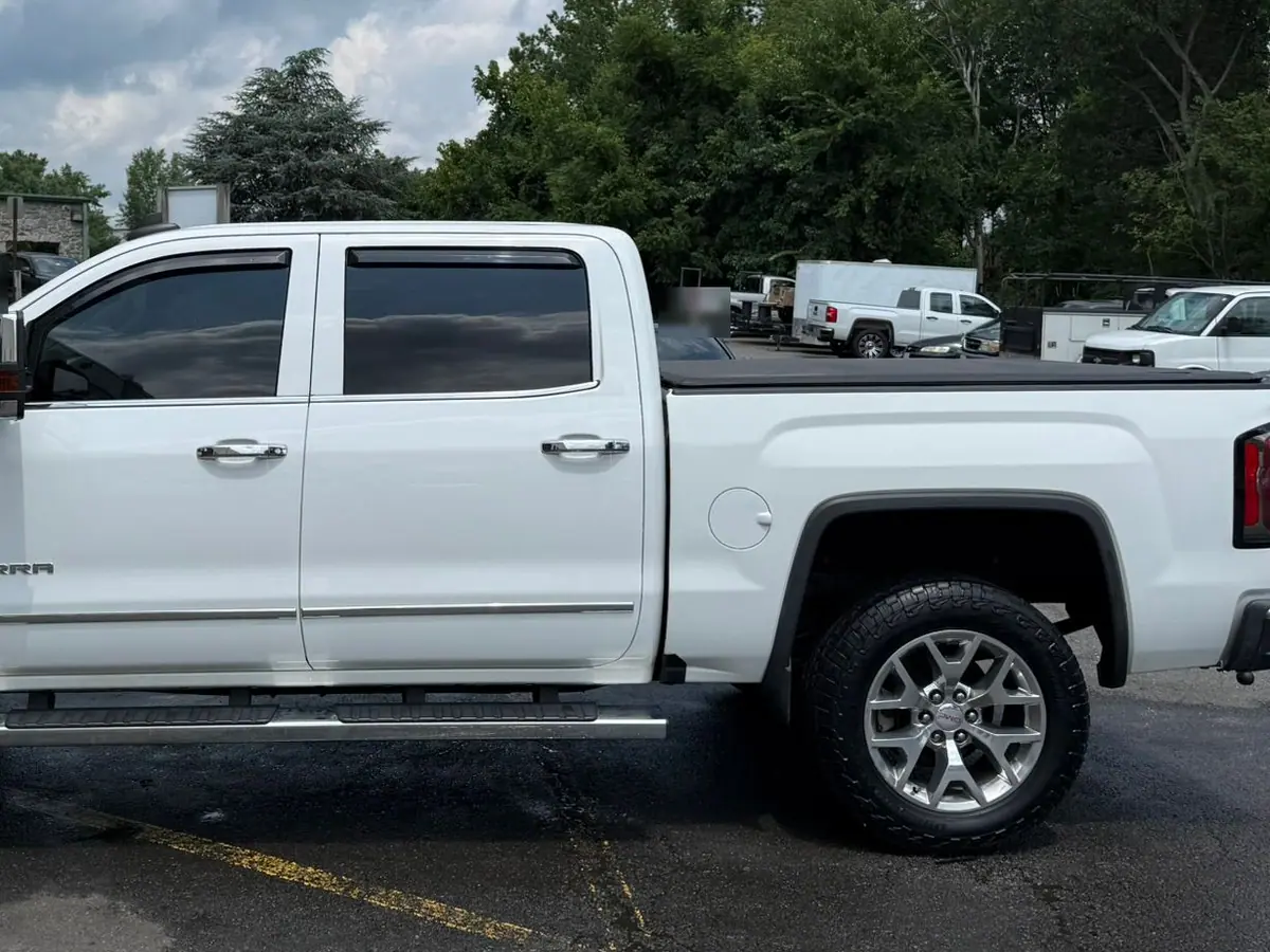 A white GMC Sierra crew cab pickup truck parked side-on across an asphalt forecourt outside a light-grey brick unit with an open overhead door, under a blue sky with tall white clouds.