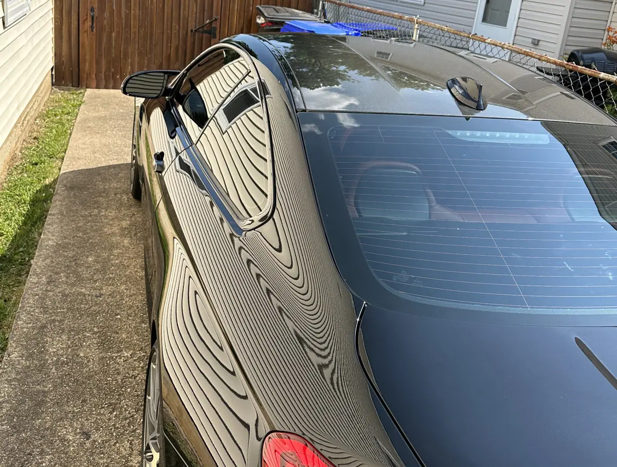 A dark grey four-door coupe parked nose-in on a driveway between two suburban houses, seen from above and behind with neighboring siding reflected across its paint.