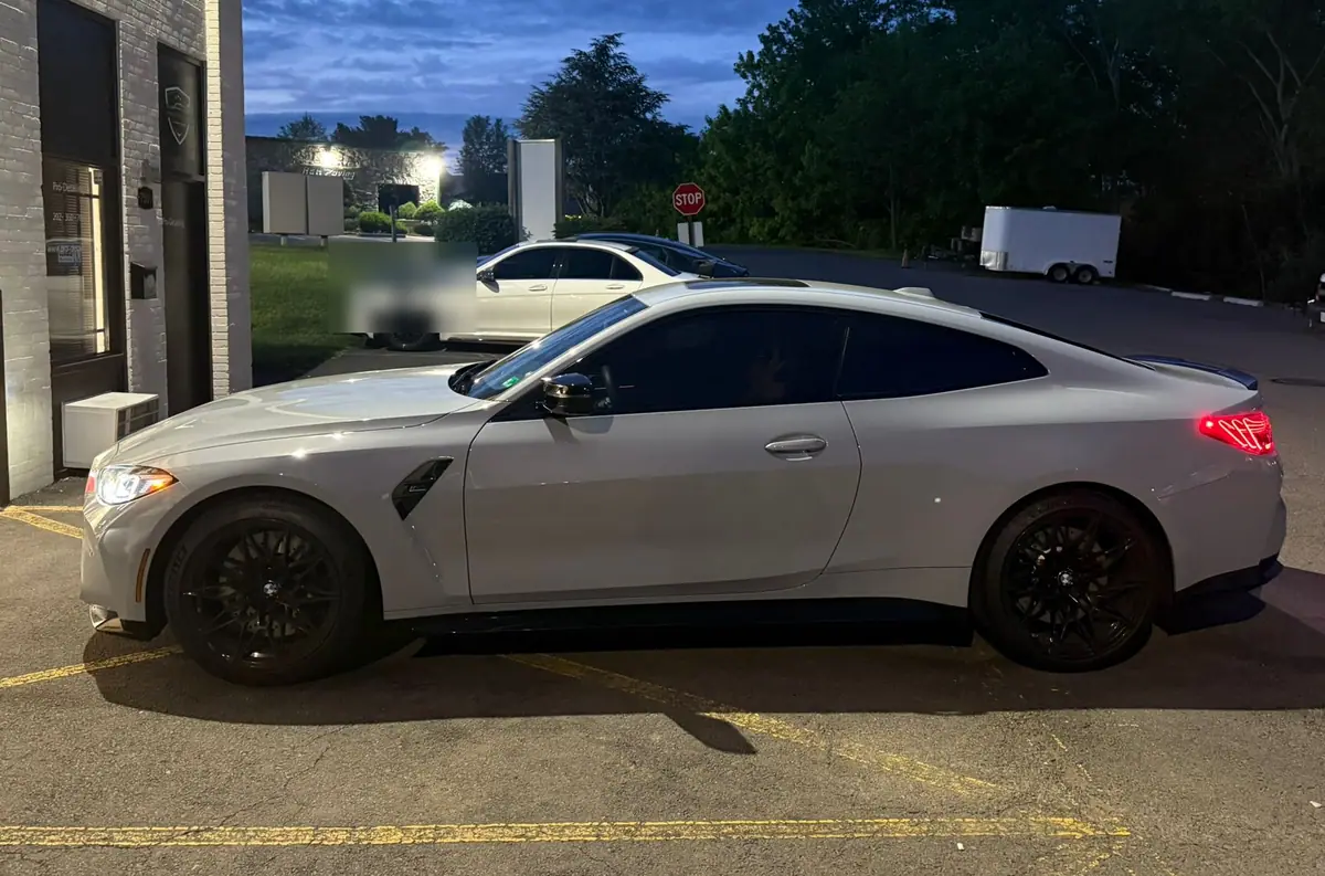 A grey BMW M4 coupe parked in side profile on the lot at dusk, its tail lights lit against a deep blue sky.