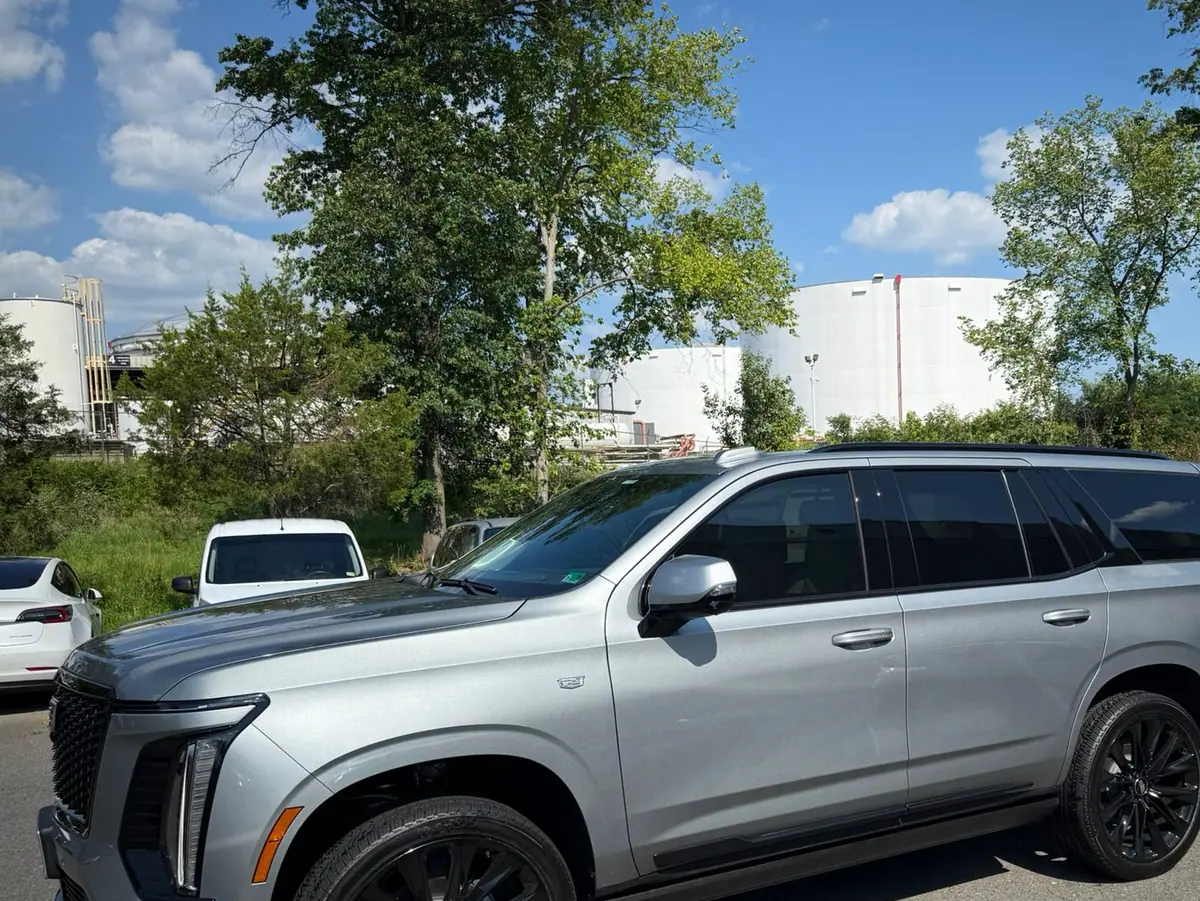 A silver Cadillac Escalade parked in side profile on the forecourt, with industrial storage tanks and summer trees behind it. PRO Detailing’s Manassas studio, serving Centreville, VA drivers.