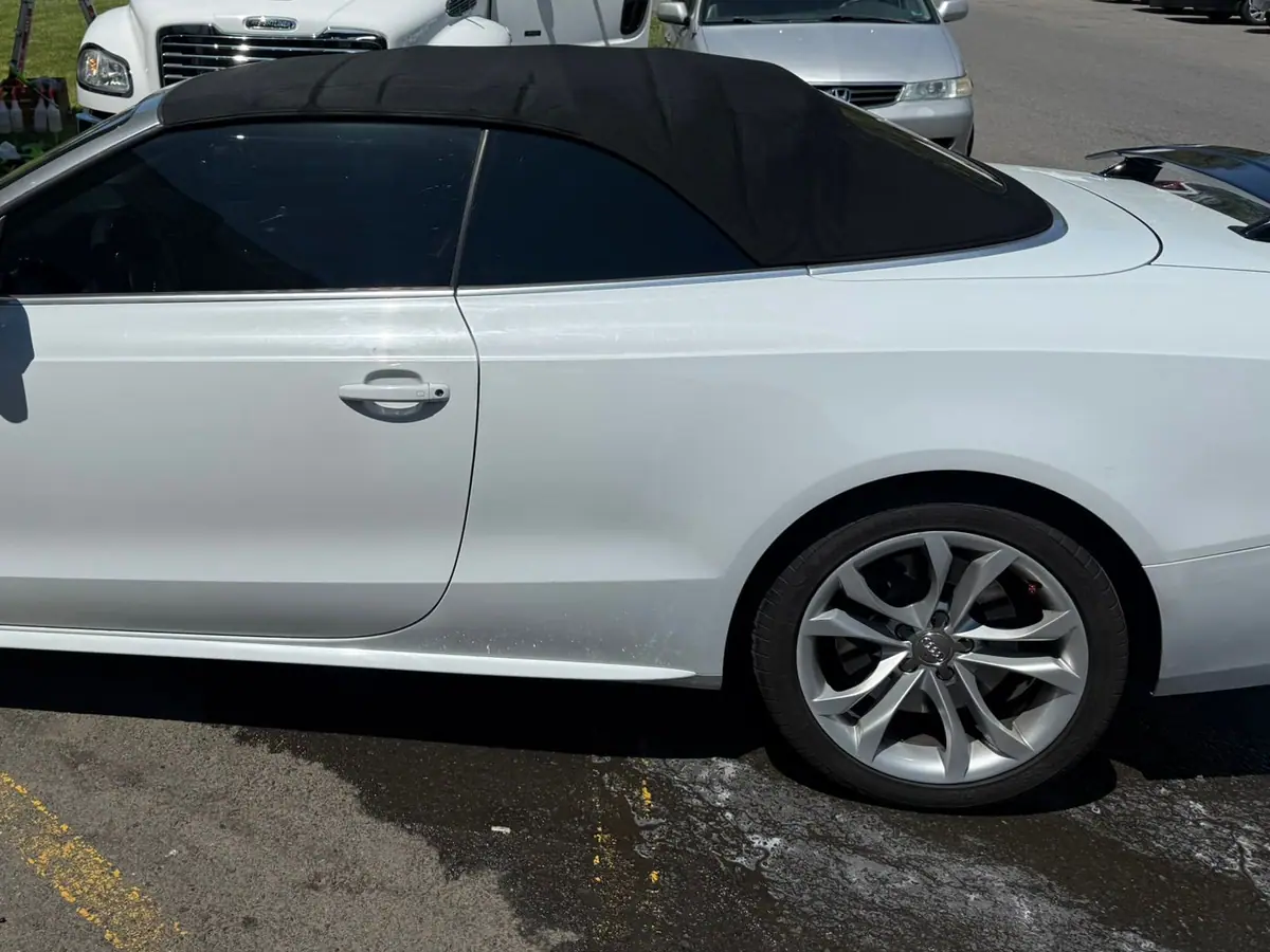 A white Audi convertible with its black fabric roof raised, parked in side profile outside a white brick unit, with a white box truck and silver minivan behind it.