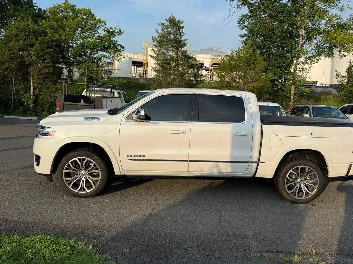A white RAM 1500 Limited crew-cab pickup parked side-on across an asphalt forecourt, with trees and industrial storage tanks behind it. PRO Detailing’s Manassas studio, serving Haymarket, VA drivers.