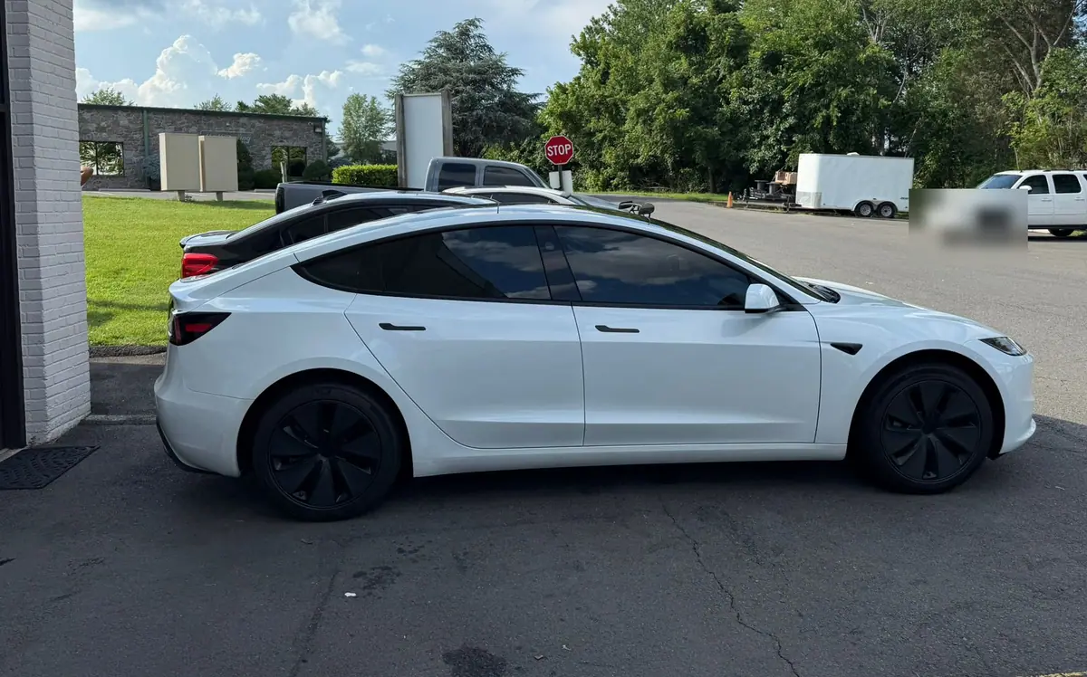 A white Tesla Model 3 parked in side profile beside the studio’s brick wall, with a stop sign and parked vehicles further down the lot. PRO Detailing’s Manassas studio, serving Chantilly, VA drivers.